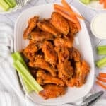 A white platter of saucy baked chicken wings is garnished with fresh celery and carrot sticks. A bowl of ranch dressing and more veggies are visible nearby on a light patterned surface.