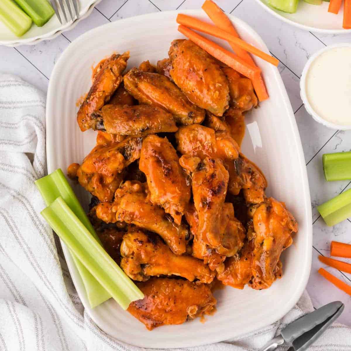 A white platter of saucy baked chicken wings is garnished with fresh celery and carrot sticks. A bowl of ranch dressing and more veggies are visible nearby on a light patterned surface.