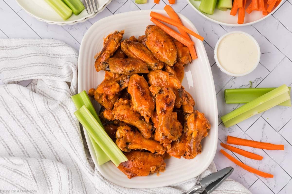 A platter of baked chicken wings is surrounded by celery and carrot sticks, with a bowl of ranch dressing nearby. The food is arranged on a white table with a striped towel and serving tongs.
