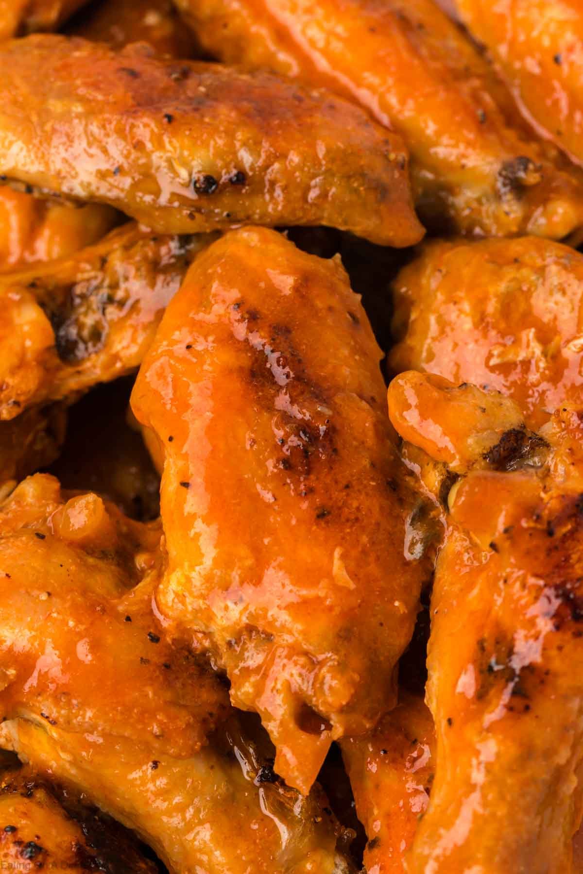 A close-up of several golden-brown baked chicken wings coated in a shiny, orange buffalo sauce. The wings appear juicy and well-seasoned, with visible grill marks and a glossy finish.