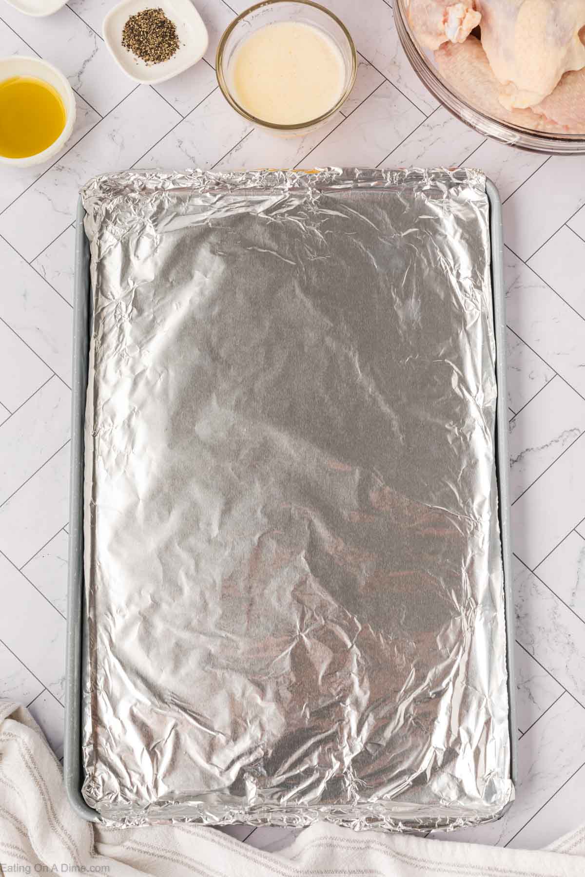 A baking sheet covered with aluminum foil sits on a white tile surface, surrounded by small bowls containing spices, a creamy liquid, and raw chicken wings ready to be transformed into delicious baked chicken wings.