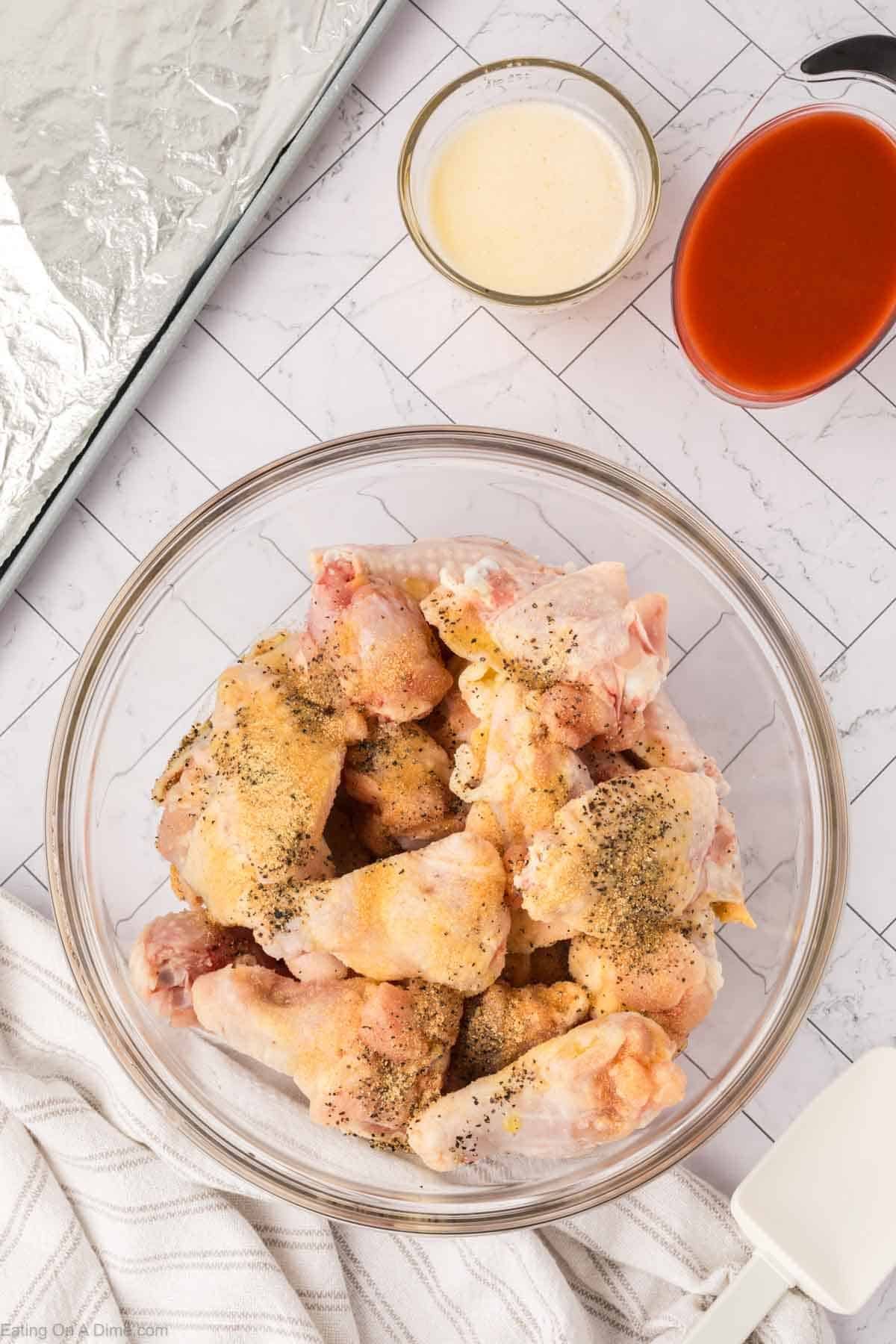 A glass bowl filled with raw, seasoned chicken wings sits on a white tile surface next to small bowls of creamy sauce and red hot sauce, a striped cloth, and a foil-covered baking sheet ready for making delicious baked chicken wings.