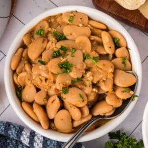 A white bowl filled with a delicious butter beans recipe, garnished with chopped parsley, with a spoon resting inside. The bowl sits on a white tiled surface next to a blue cloth and a wooden board.