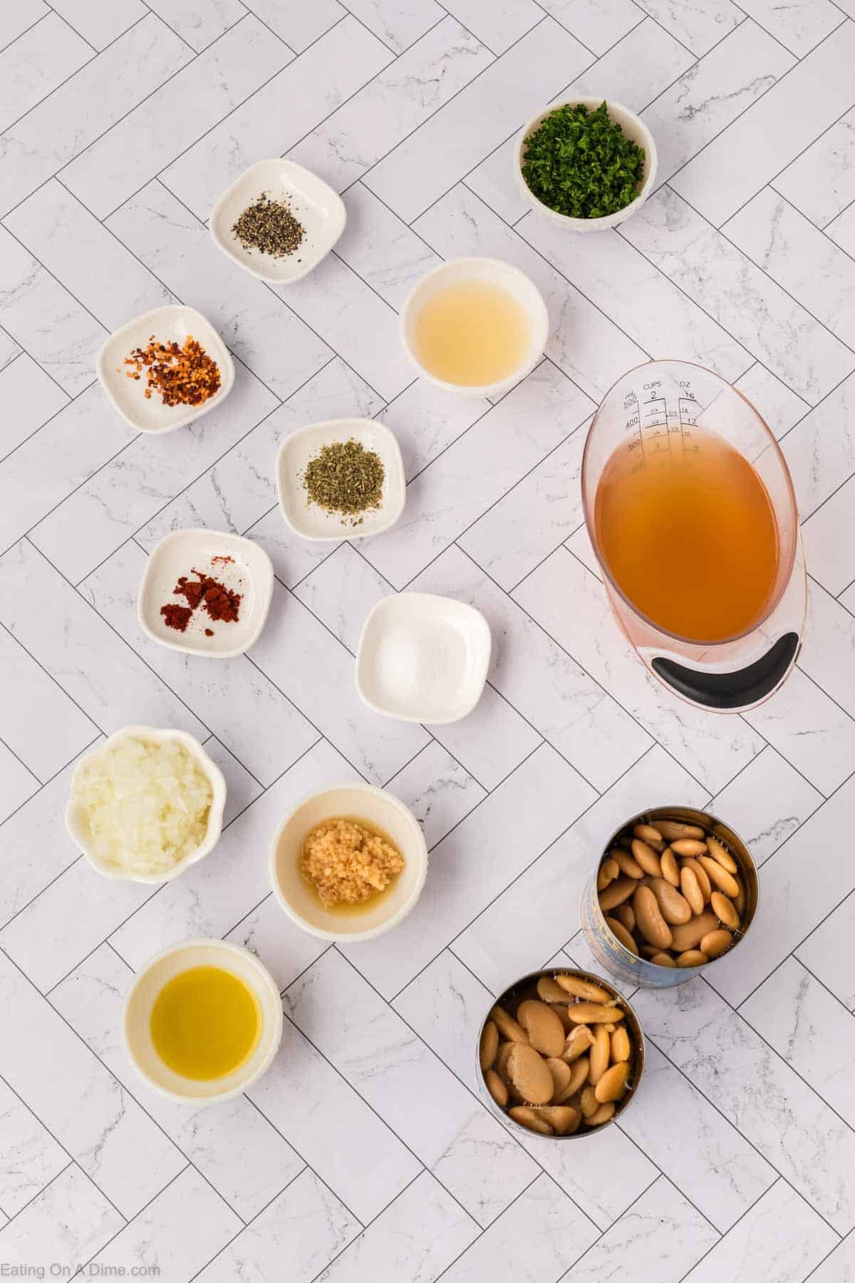 A top-down view of various ingredients in small bowls on a white tiled surface, perfect for a Butter Beans Recipe, including seasonings, broth, chopped herbs, minced garlic, olive oil, onions, and two open cans of white beans.