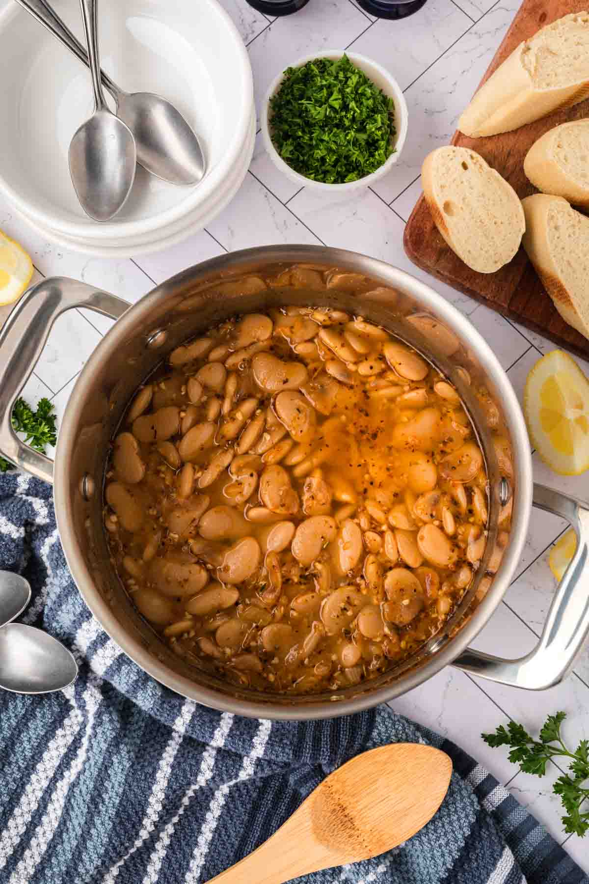 A pot of cooked beans from a classic butter beans recipe in seasoned broth sits on a striped towel, surrounded by sliced bread, chopped parsley, lemon wedges, stacked white bowls, and spoons on a white countertop.