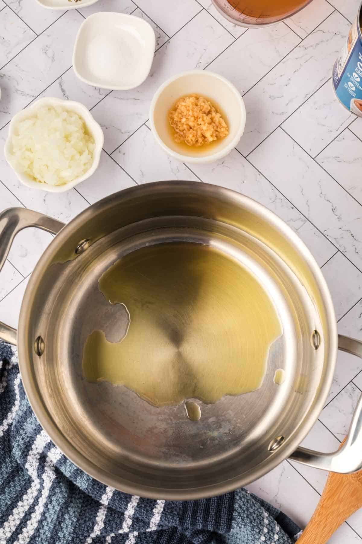 A stainless steel pot with oil inside on a tiled countertop, surrounded by small bowls of chopped onions, minced garlic, and salt—perfect for starting a Butter Beans Recipe. A blue-striped towel is partially visible in the corner.