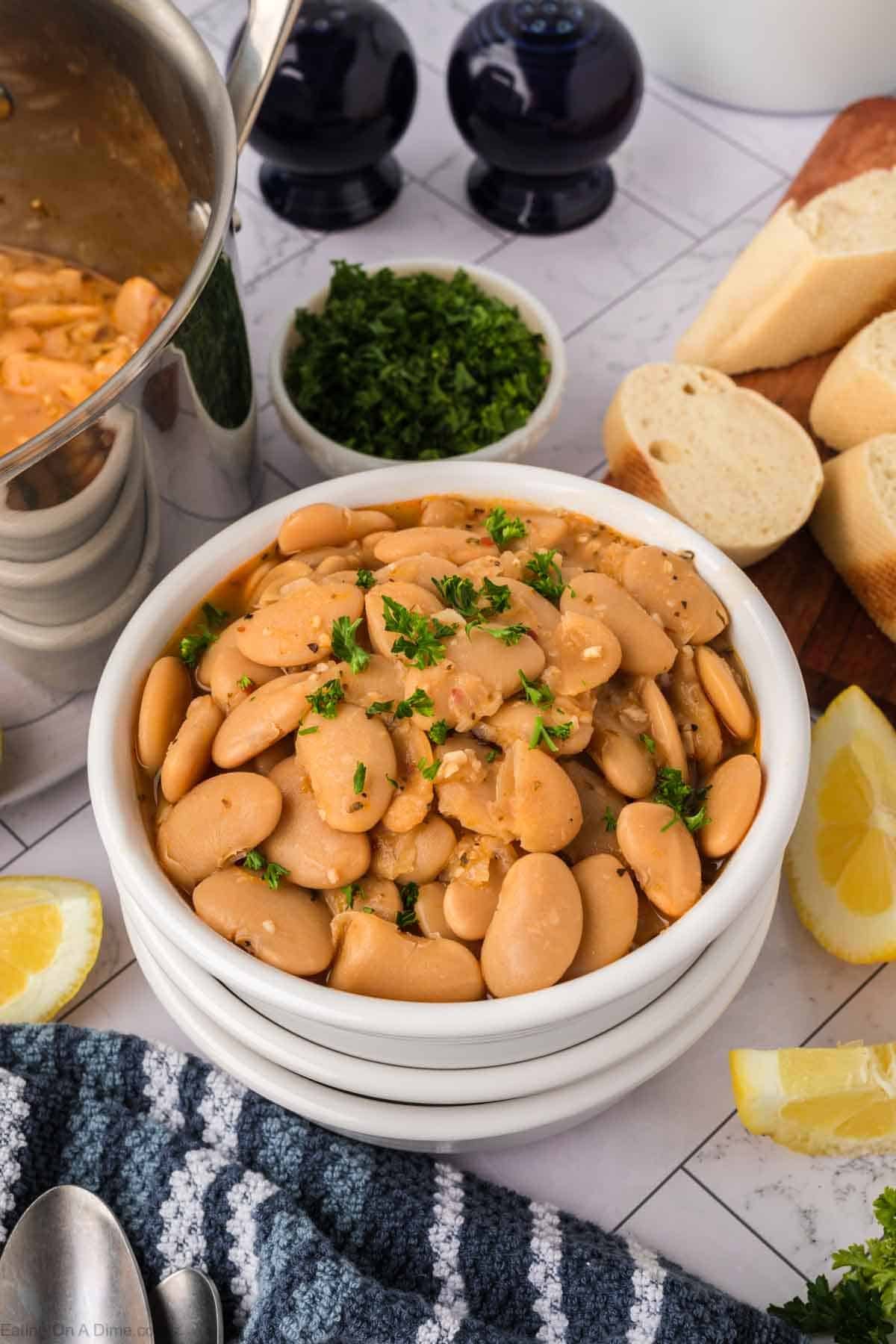 A white bowl filled with cooked butter beans is garnished with chopped parsley, showcasing a classic Butter Beans Recipe. It's surrounded by lemon wedges, sliced bread, fresh herbs, and a pot of beans on a tiled surface with a blue-striped towel nearby.