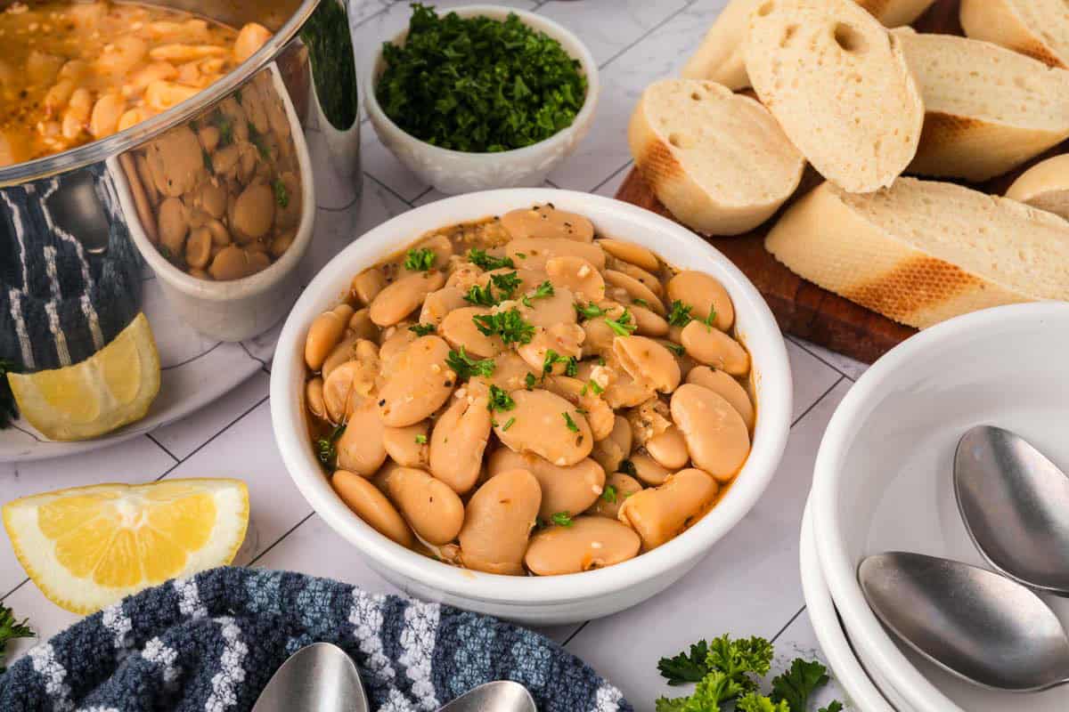 A bowl of cooked butter beans garnished with chopped parsley sits on a table beside sliced baguette, a pot of beans, a lemon wedge, chopped herbs, and stainless steel spoons—an inviting Butter Beans Recipe to savor.