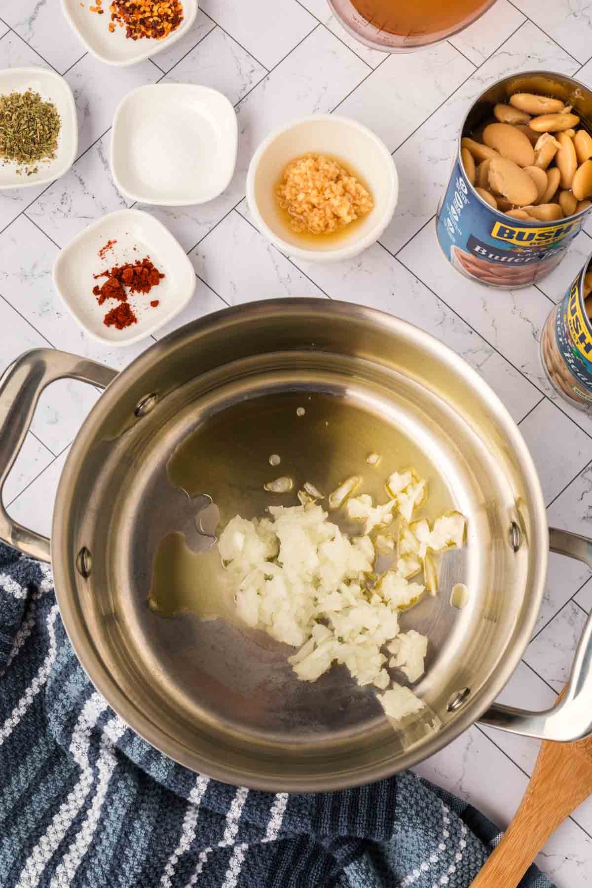 A metal pot on a white tile surface contains chopped onions sautéing in oil. Nearby are small bowls of spices, minced garlic, a can of white beans for your Butter Beans Recipe, a striped cloth, and a wooden spoon.
