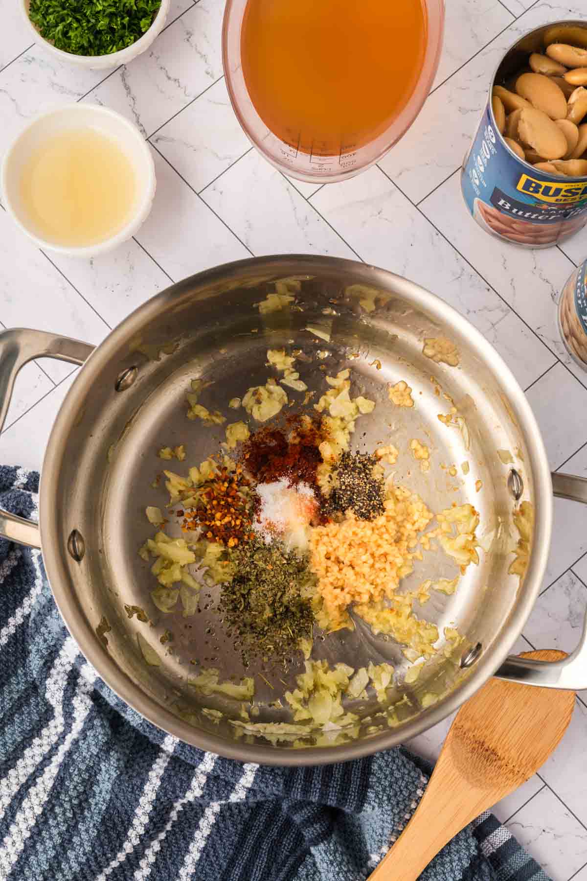 A stainless steel pot on a white countertop contains sautéed onions, minced garlic, and assorted herbs and spices. Surrounding the pot for this Butter Beans Recipe are a striped towel, wooden spoon, broth, canned beans, lemon juice, and chopped herbs.