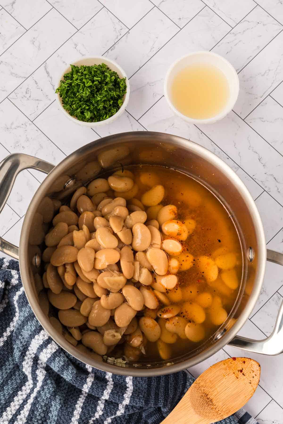 A pot of simmering butter beans sits in broth with spices on a blue-striped towel. Nearby are bowls of fresh herbs and a light liquid, perfect for finishing this butter beans recipe. A wooden spoon rests beside the hearty dish.