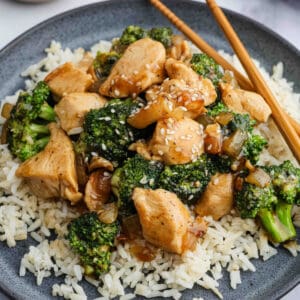 A plate of white rice topped with Chicken Broccoli Stir Fry featuring tender chicken, broccoli, onions, and sesame seeds, served with wooden chopsticks.