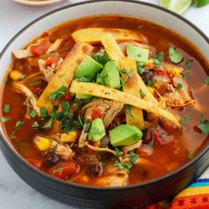 A bowl of Chicken Tortilla Soup with shredded chicken, diced avocado, tortilla strips, corn, black beans, and diced tomatoes, garnished with fresh cilantro. The soup has a rich red color and is served with lime and colorful fabric underneath.