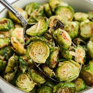 A close-up of Parmesan Brussel Sprouts, roasted to perfection and topped with grated cheese, served in a bowl with a spoon lifting a portion.