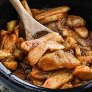 A wooden spoon stirs sliced apples coated in a cinnamon sugar sauce inside a black pot, creating delicious Slow Cooker Apples reminiscent of classic apple pie filling or a warm dessert.