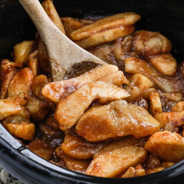 A wooden spoon stirs sliced apples coated in a cinnamon sugar sauce inside a black pot, creating delicious Slow Cooker Apples reminiscent of classic apple pie filling or a warm dessert.