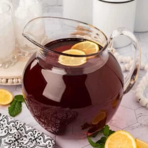 A glass pitcher filled with iced tea, inspired by a classic sweet tea recipe and garnished with lemon slices, sits on a marble countertop. Mint leaves, extra lemon slices, and a patterned napkin are arranged nearby. Two glasses with ice are in the background.