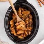 A wooden spoon stirs sliced apples coated in cinnamon and brown sugar inside a black slow cooker, creating delicious Slow Cooker Apples on a light countertop.