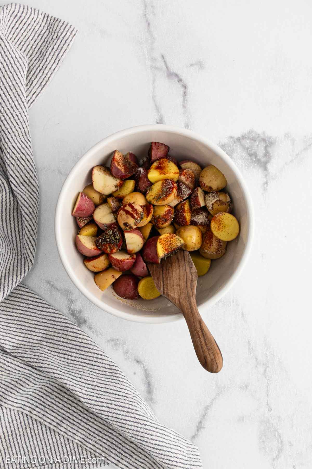 A white bowl filled with seasoned, chopped red and yellow potatoesโperfect for making crispy Air Fryer Potatoesโrests on a white marble surface next to a gray-striped cloth, with a wooden spoon inside.