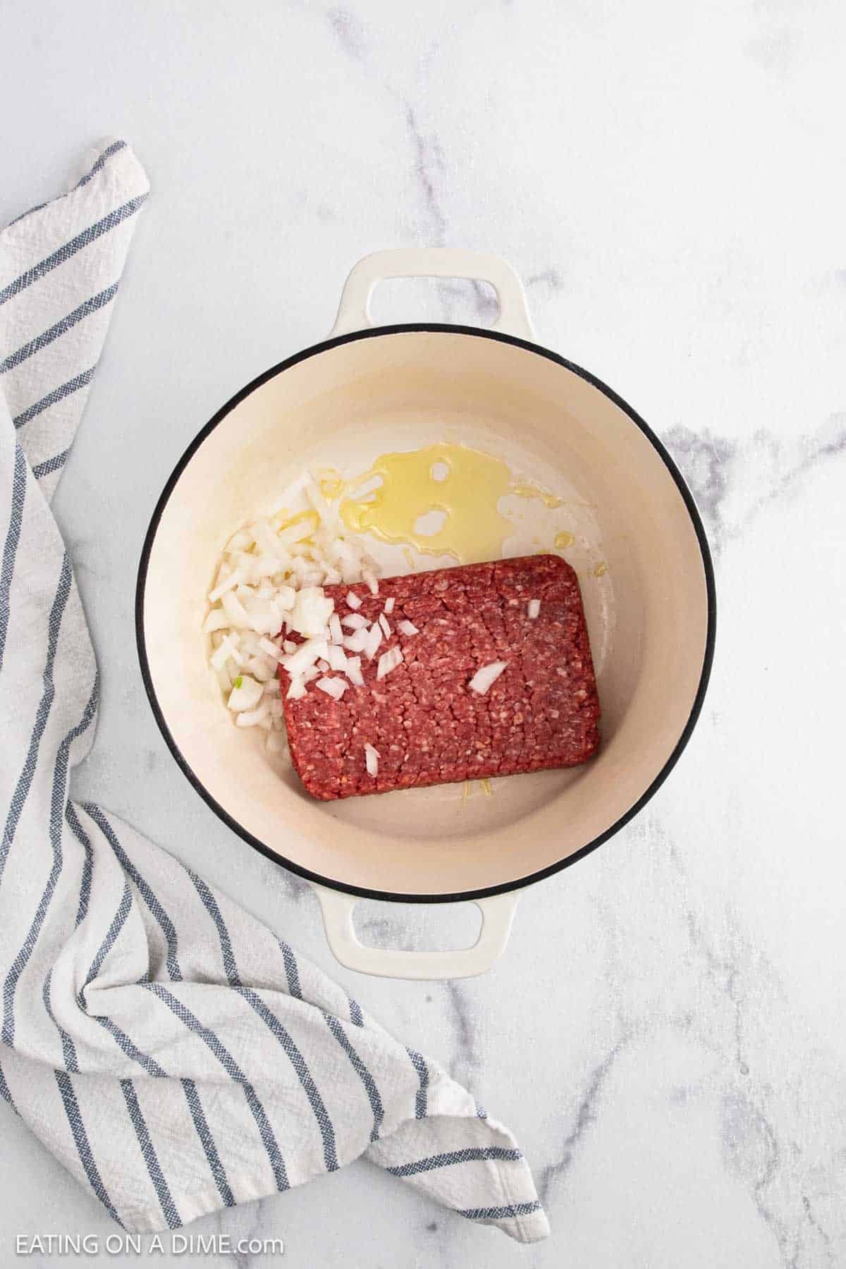 A white pot holds raw ground beef, chopped onions, and oilโperfect beginnings for a hearty Stuffed Cabbage Soup. The pot sits on a white marble surface next to a striped kitchen towel, ready for the next step.