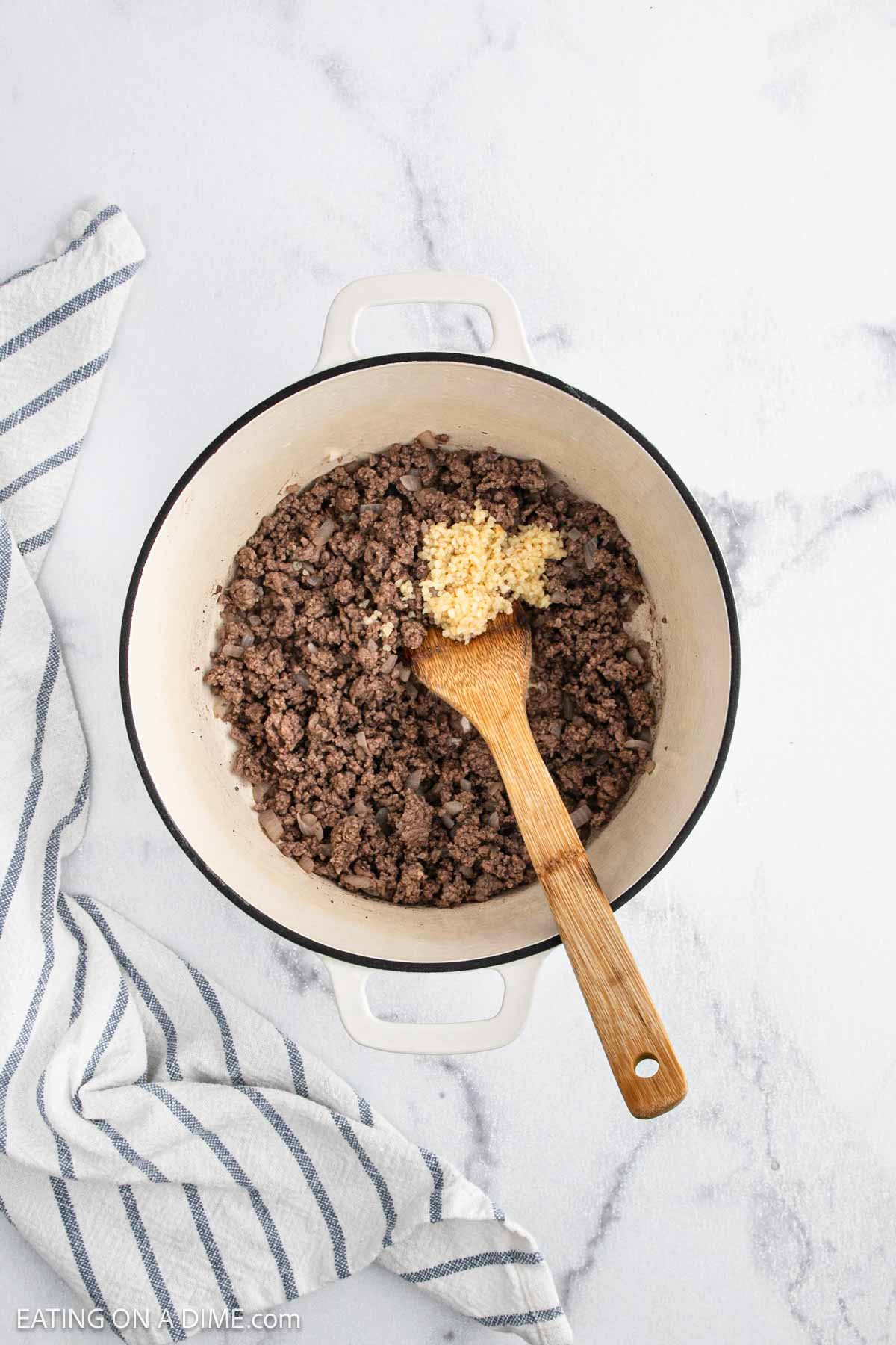 A white pot with cooked ground beef and minced garlic is being stirred by a wooden spoon, as the base for hearty Cabbage Roll Soup, on a white marble surface next to a striped kitchen towel.