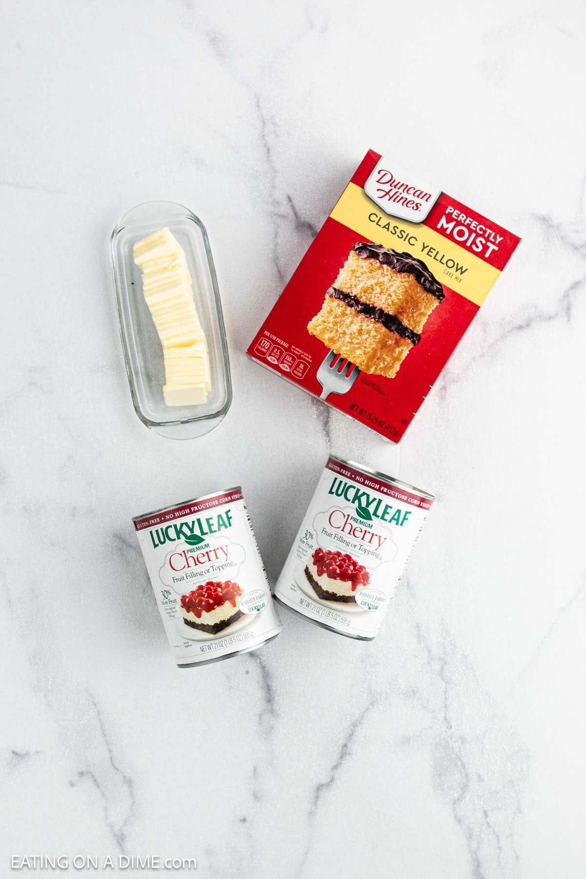 A glass dish with sliced butter, a box of Duncan Hines Classic Yellow cake mix, and two cans of Lucky Leaf cherry pie filling on a white marble surface—perfect for making an easy Cherry Dump Cake.