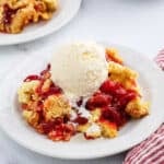 A plate of cherry cobbler topped with a scoop of vanilla ice cream, melting down the sides. Another plate of cherry dump cake is in the background, along with a red and white striped napkin.