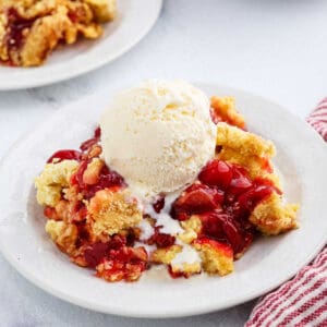 A plate of cherry cobbler topped with a scoop of vanilla ice cream, melting down the sides. Another plate of cherry dump cake is in the background, along with a red and white striped napkin.
