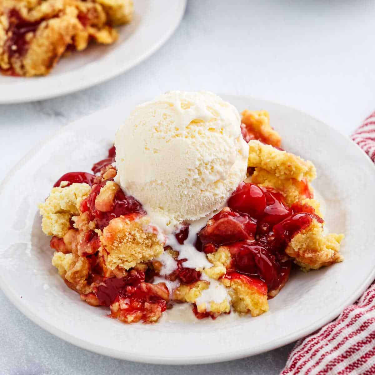 A plate of cherry cobbler topped with a scoop of vanilla ice cream, melting down the sides. Another plate of cherry dump cake is in the background, along with a red and white striped napkin.
