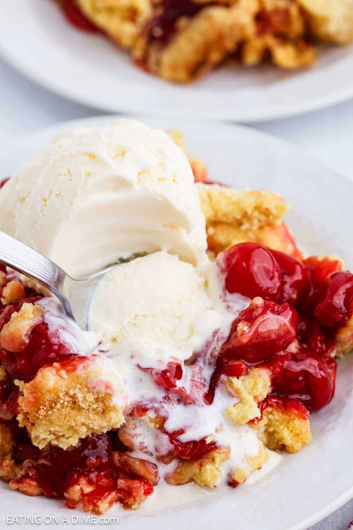 A close-up of a dessert plate featuring Cherry Dump Cake topped with a melting scoop of vanilla ice cream. A spoon is scooping up some of the ice cream and cake, revealing juicy cherries and crumbly topping.