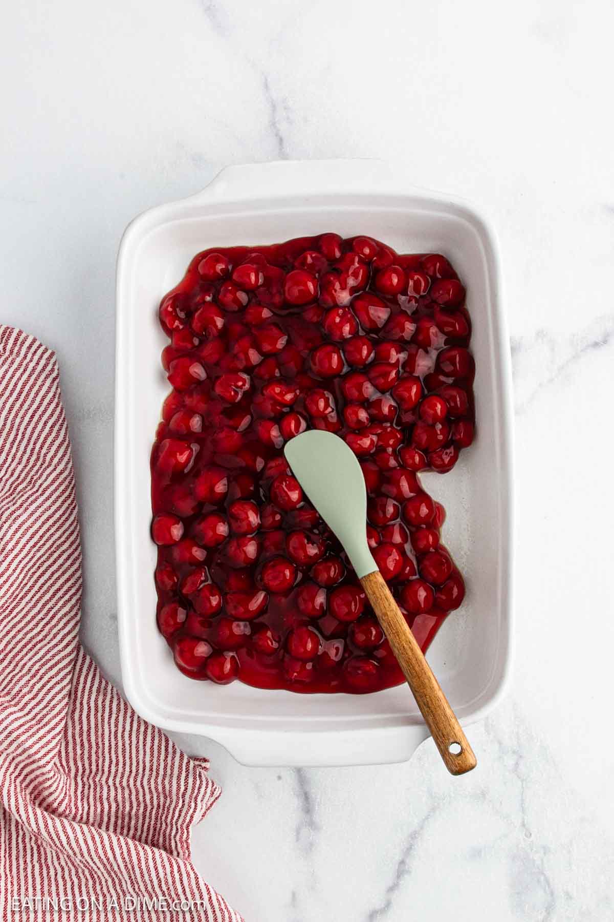 A white baking dish filled with cherry dump cake filling and a green and wooden spoon resting inside. A red and white striped kitchen towel is placed beside the dish on a white marble surface.