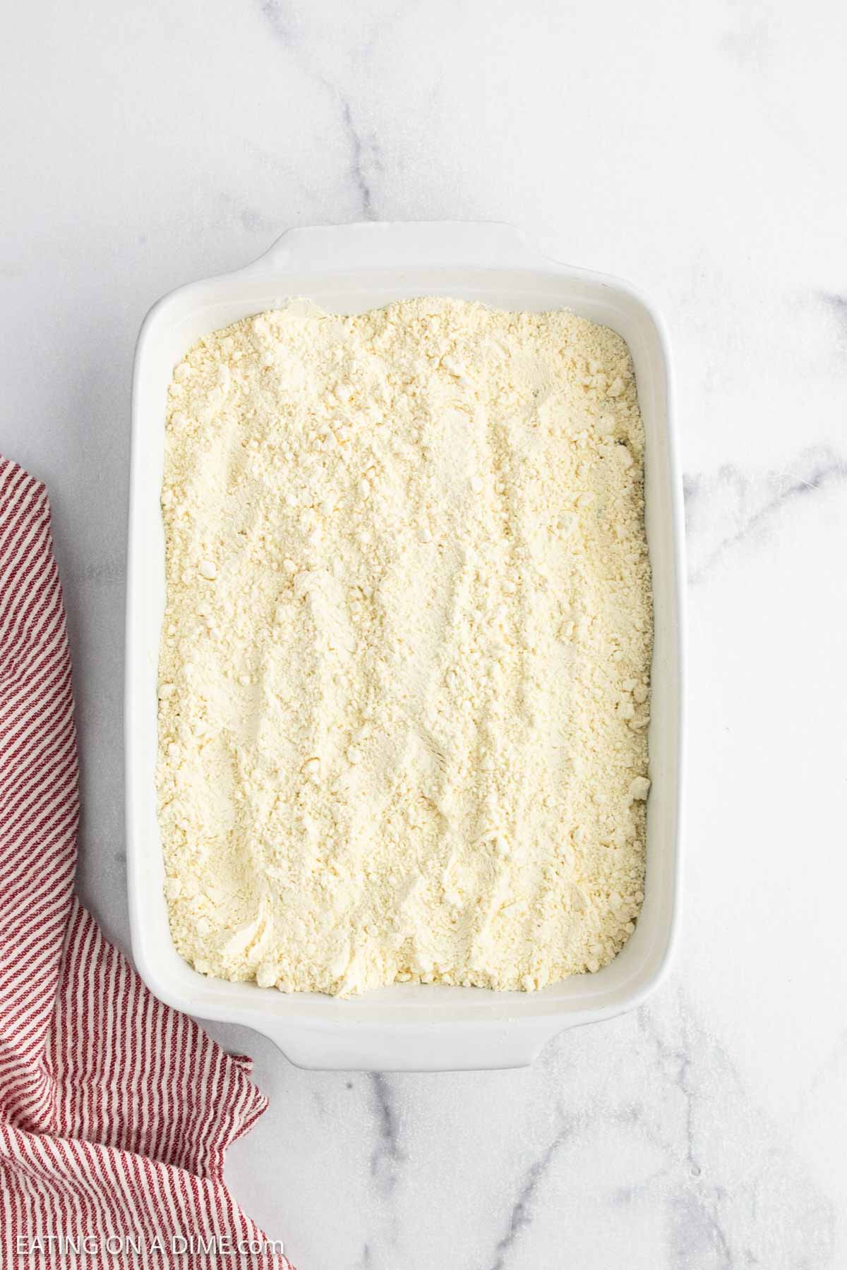 A white rectangular baking dish filled with a layer of dry, crumbly biscuit mix for Cherry Dump Cake sits on a white marble surface beside a red and white striped cloth.