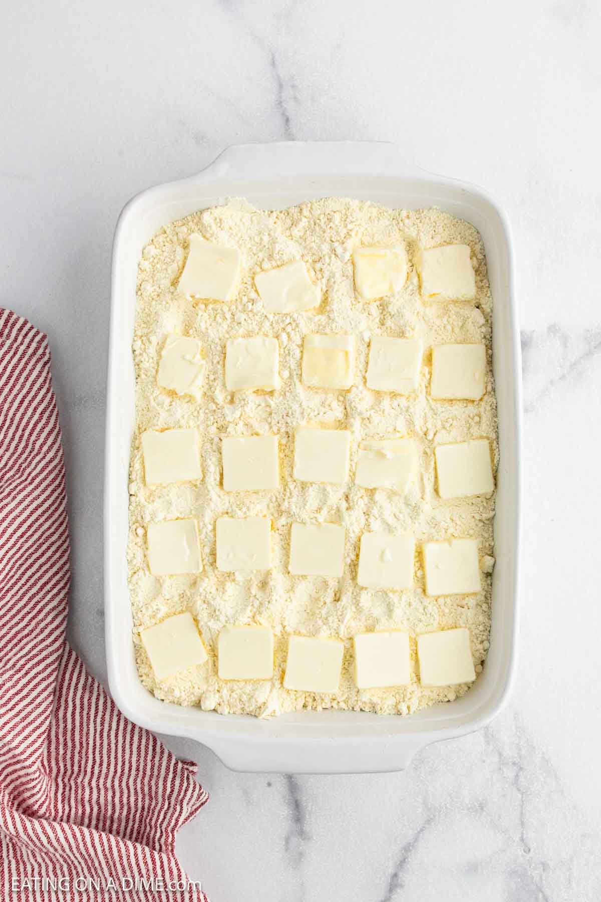 A white baking dish filled with crumbly Cherry Dump Cake mixture is topped with evenly spaced square slices of butter. The dish sits on a white marble surface next to a red and white striped kitchen towel.