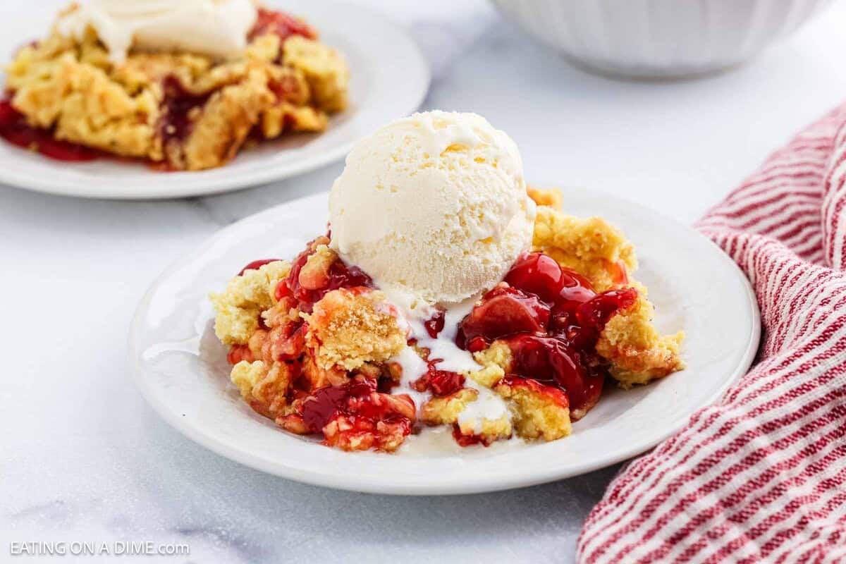 A plate of cherry cobbler, inspired by a classic Cherry Dump Cake recipe, is topped with melting vanilla ice cream on a white plate beside a red and white striped cloth. Another cherry dessert sits blurred in the background.