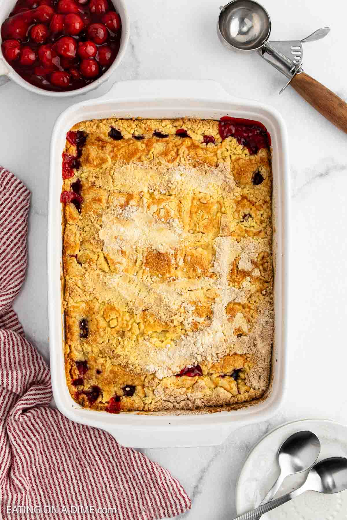 A baked Cherry Dump Cake in a white casserole dish sits on a marble countertop, with a scoop, two spoons, a red and white striped towel, and a bowl of cherry filling nearby—an irresistible cherry dessert display.
