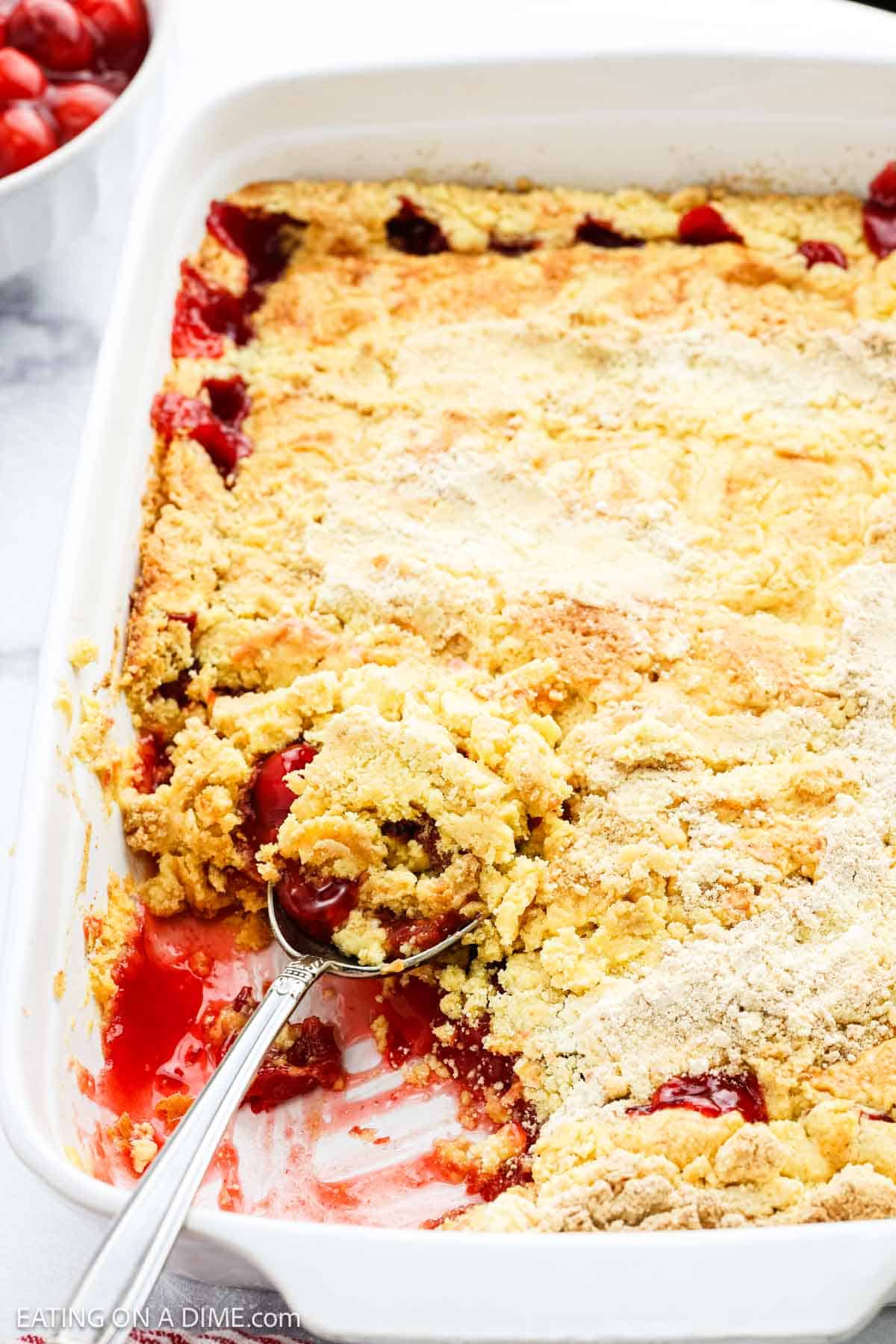 A close-up of a Cherry Dump Cake in a white baking dish, with a serving spoon scooping out a portion. The cake has a golden, crumbly top and visible red cherry filling underneath, making this cherry dessert look irresistible.