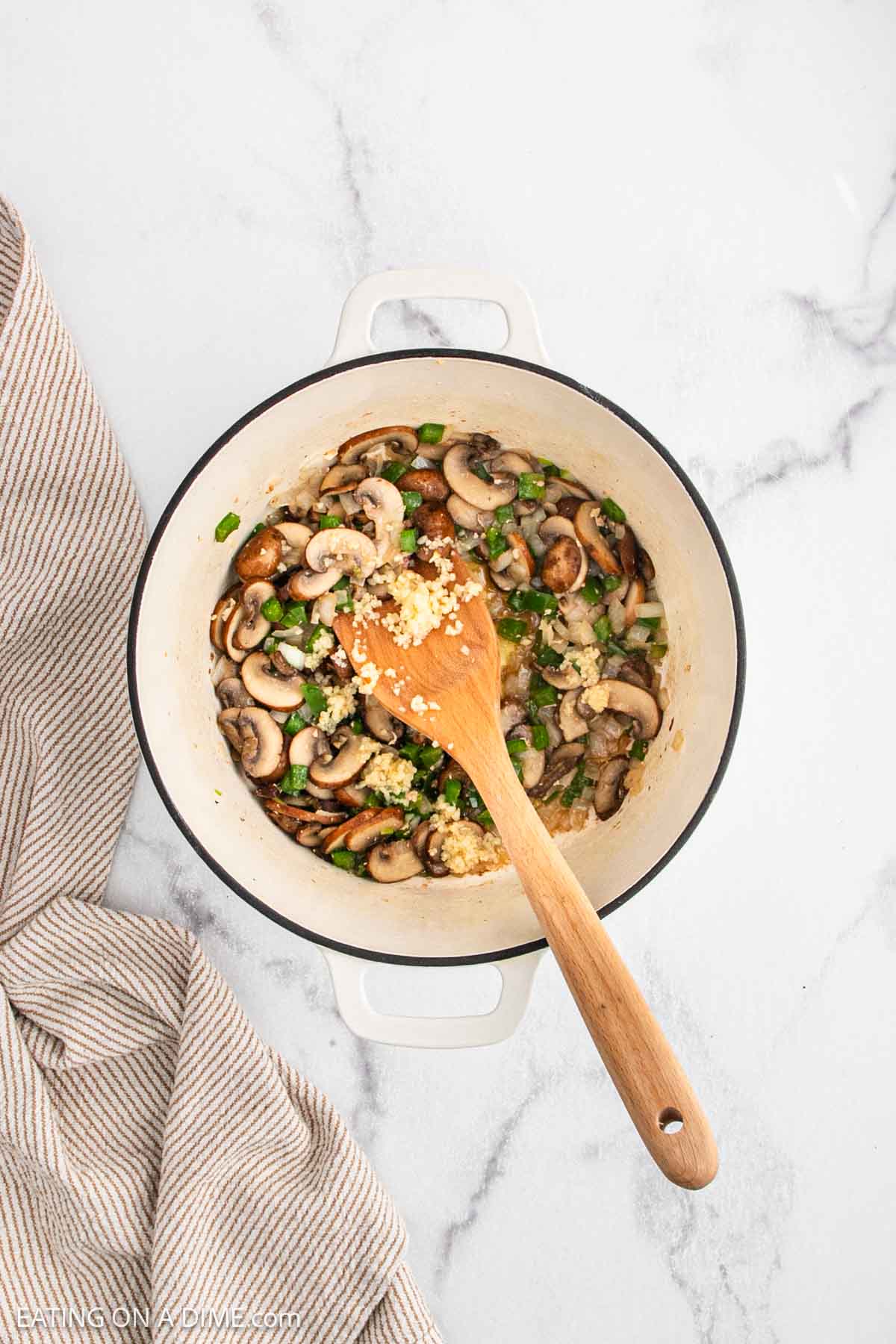A white pot filled with sautรฉed mushrooms, onions, and spinachโclassic ingredients for Chicken a la Kingโis being stirred with a wooden spoon on a marble countertop beside a beige striped towel.