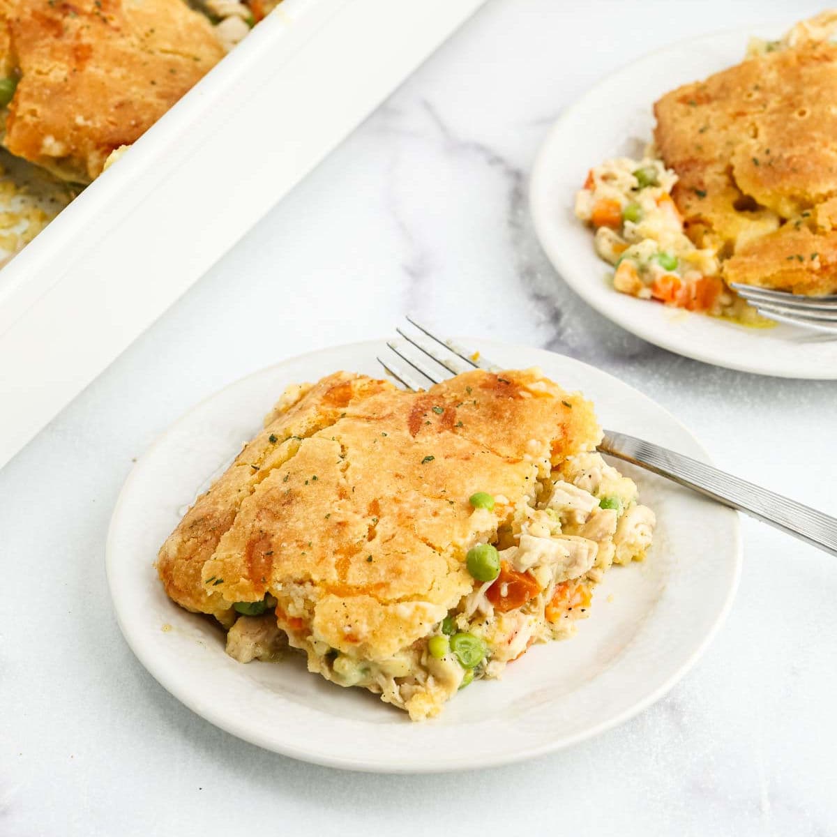 A serving of chicken pot pie with a golden biscuit crust—reminiscent of Chicken Cobbler—filled with chicken, peas, and carrots sits on a white plate with a fork. Another plate and a baking dish with more pot pie are in the background.