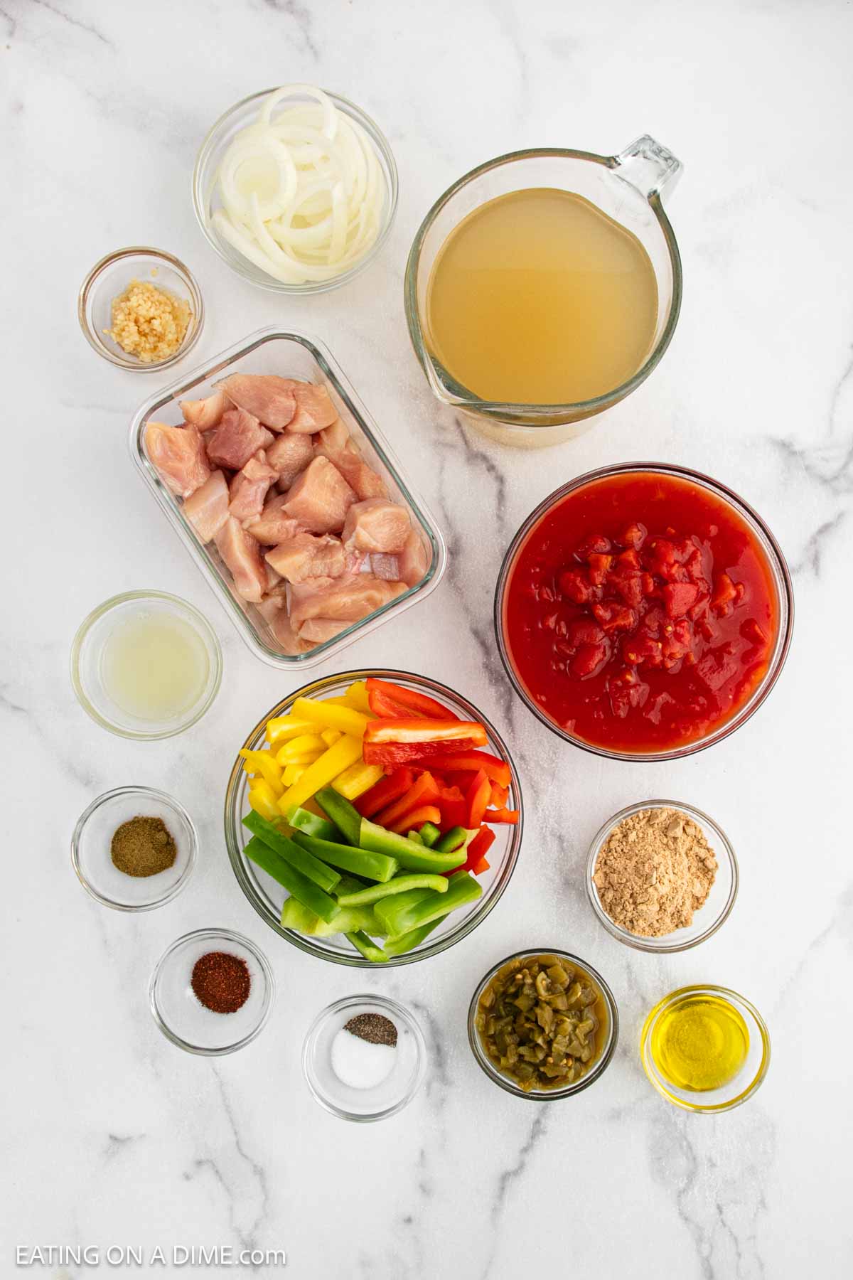Various ingredients for Chicken Fajita Soup are arranged on a white marble surface, including diced raw chicken, chopped bell peppers, diced tomatoes, chicken broth, sliced onions, seasonings, oil, minced garlic, lime juice, and chopped green chiles.
