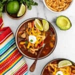 A bowl of Chicken Fajita Soup topped with shredded cheese, tortilla strips, sour cream, and lime wedges. Surrounded by avocado, more lime, extra tortilla strips, and a colorful striped napkin on a marble surface.