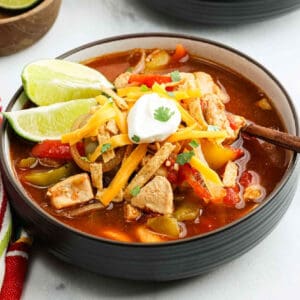 A bowl of Chicken Fajita Soup topped with shredded cheese, sour cream, cilantro, and crunchy tortilla strips, served with lime wedges on the side and a spoon in the bowl.