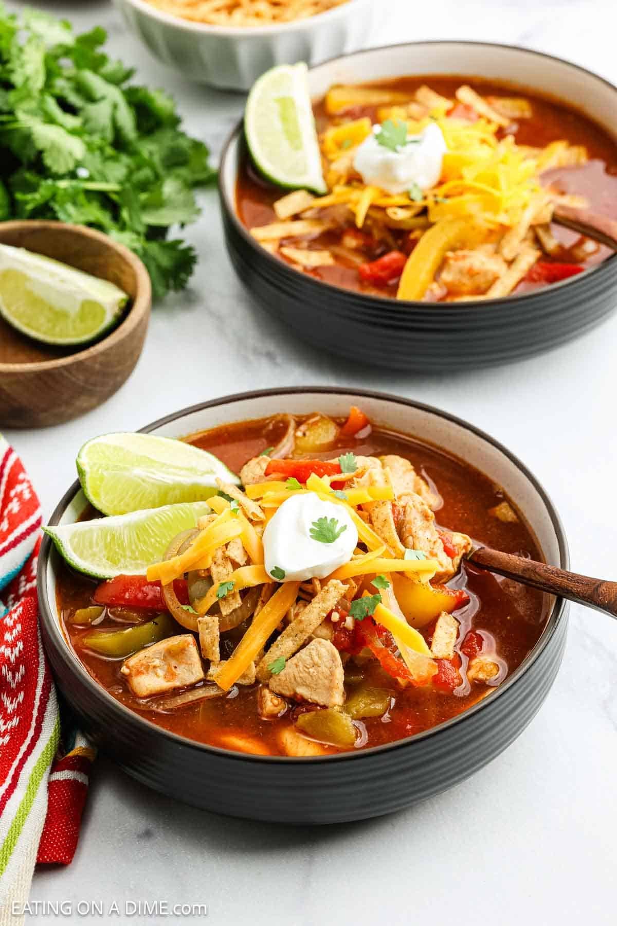 A bowl of Chicken Fajita Soup topped with shredded cheese, sour cream, tortilla strips, and cilantro, with lime wedges on the side. Another bowl of soup, lime, and cilantro are in the background on a white surface.