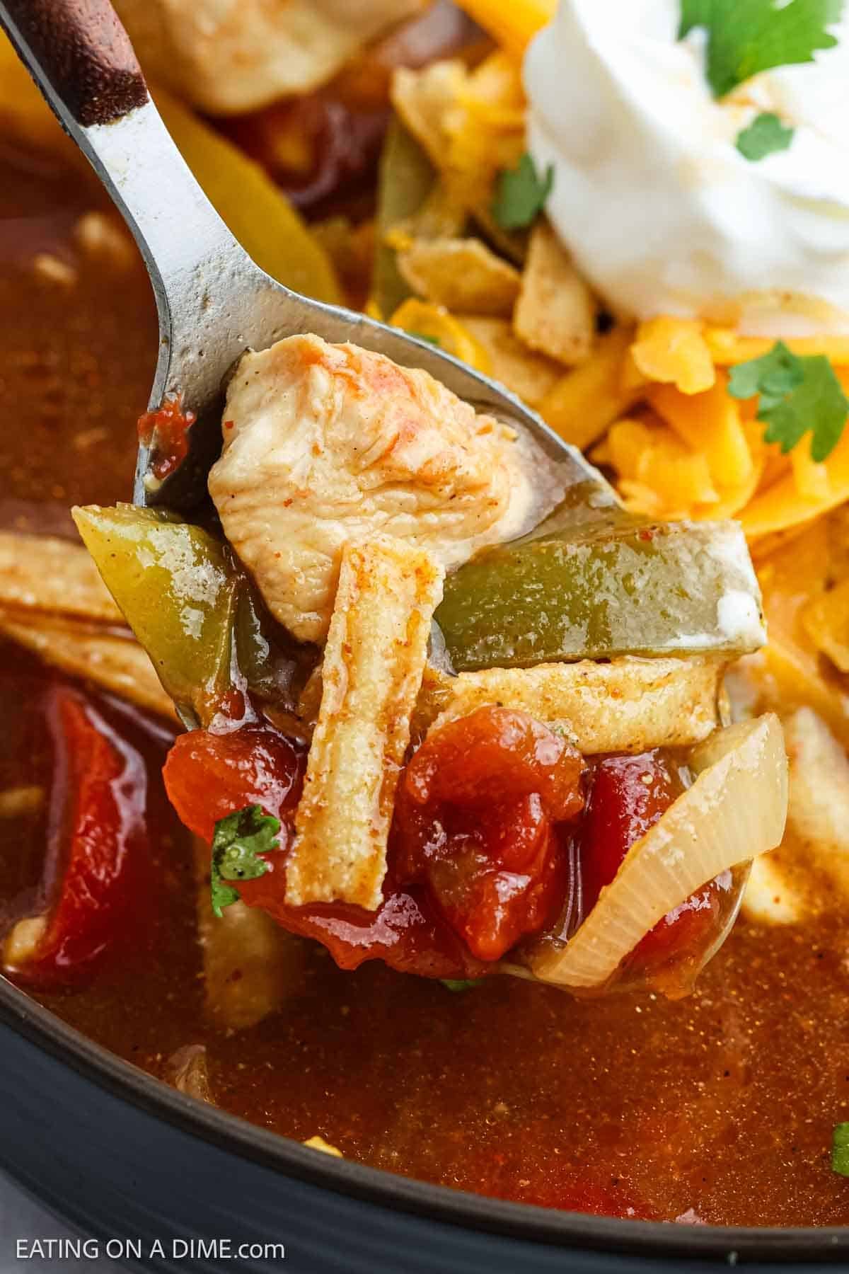 A close-up of a spoonful of Chicken Fajita Soup with chunks of chicken, tomatoes, green bell peppers, onions, tortilla strips, and shredded cheese, topped with sour cream and cilantro.