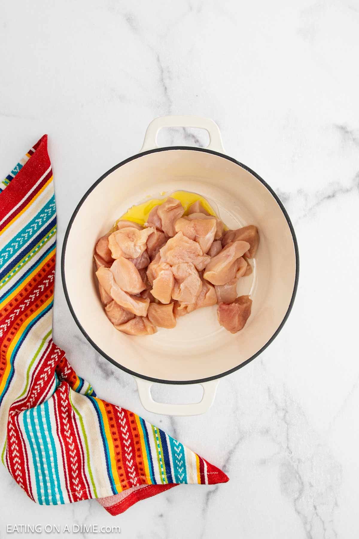 A white pot with raw chicken pieces and a bit of oil inside, ready to become Chicken Fajita Soup, sits on a white marble surface next to a colorful striped kitchen towel.
