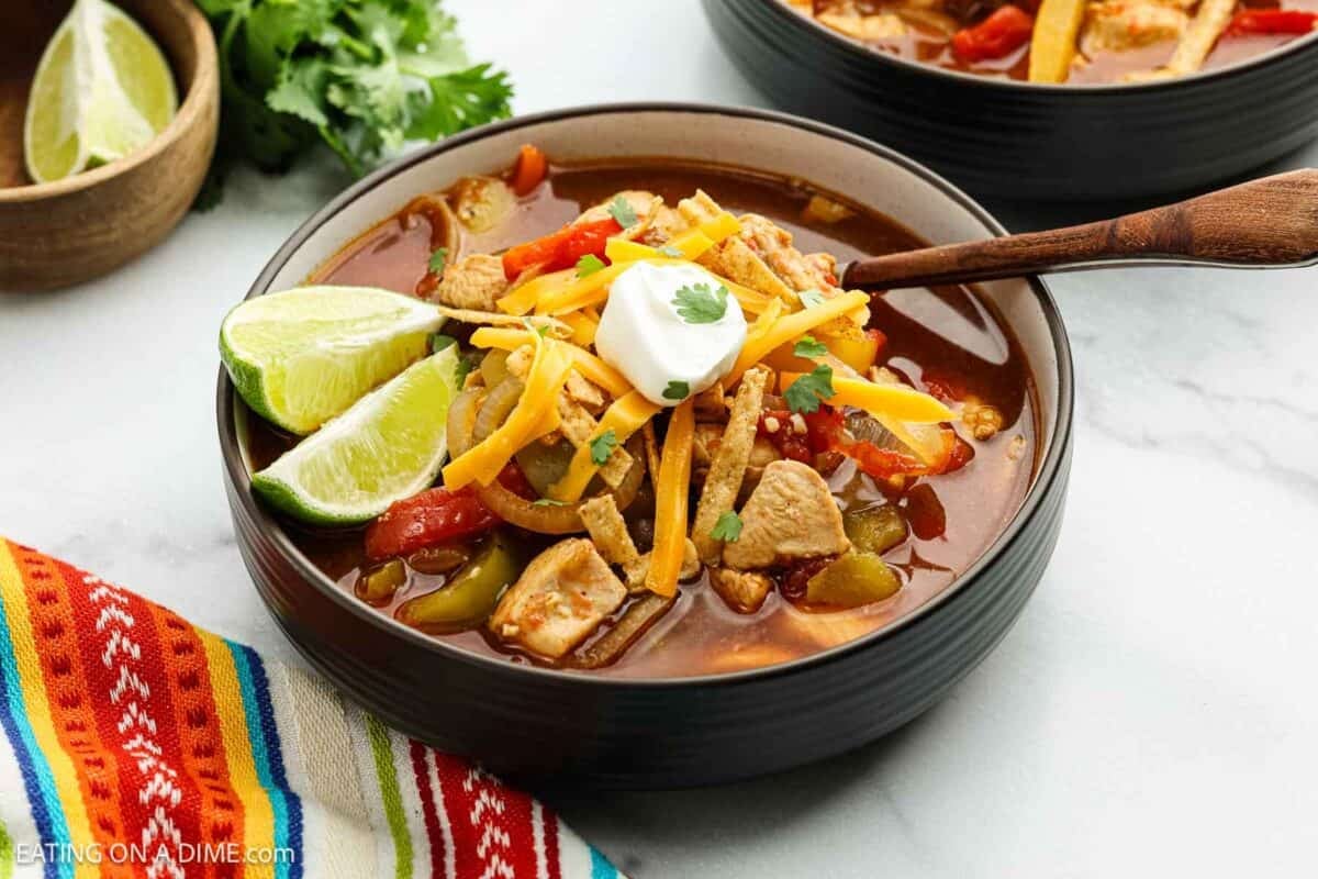 A bowl of Chicken Fajita Soup topped with shredded cheese, sour cream, tortilla strips, and a lime wedge, with a spoon in the bowl and colorful fabric nearby. Fresh cilantro and another bowl of soup are in the background.