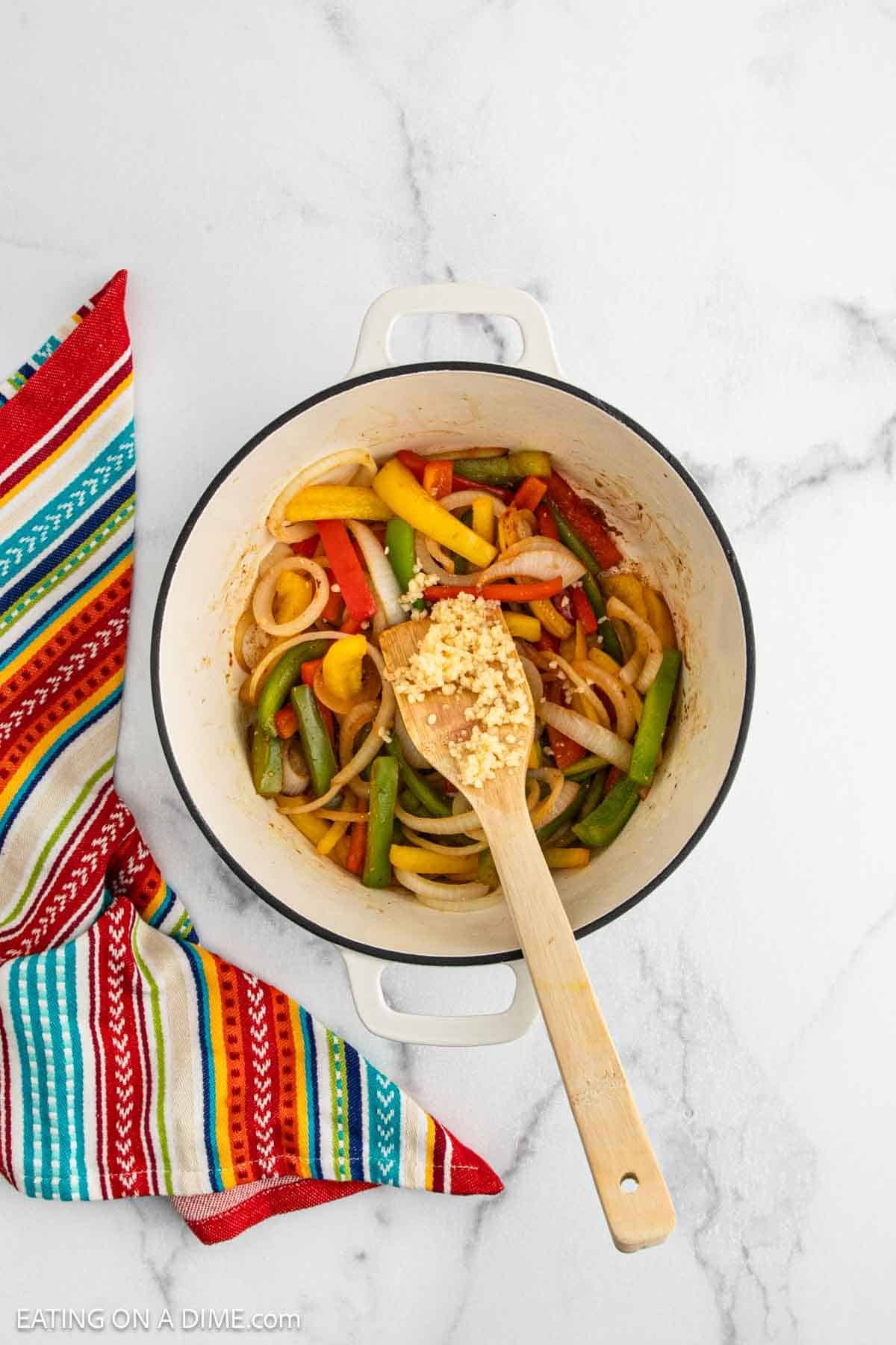 A white pot filled with sliced bell peppers, onions, and minced garlic for Chicken Fajita Soup is being stirred with a wooden spoon on a marble surface next to a colorful striped cloth.