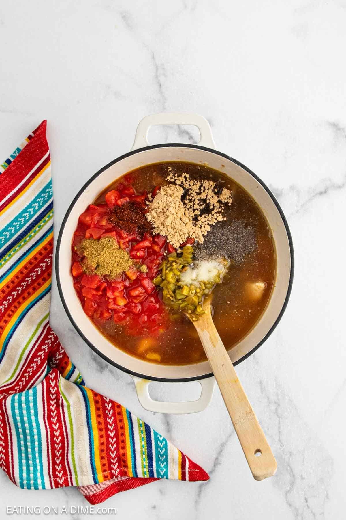A white pot of Chicken Fajita Soup with diced tomatoes, green chilies, spices, broth, and seasonings is mixed with a wooden spoon, sitting on a marble surface next to a colorful striped cloth.