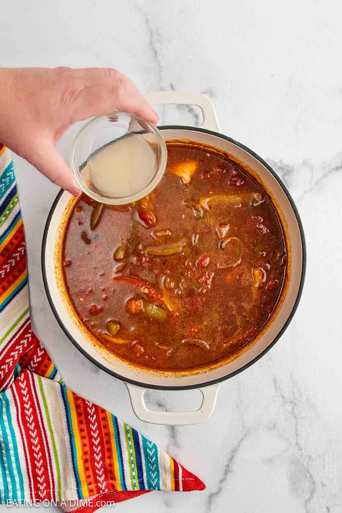A hand pours a small glass bowl of liquid into a pot of Chicken Fajita Soup filled with vegetables and beans. The pot sits on a marble surface next to a colorful striped cloth.