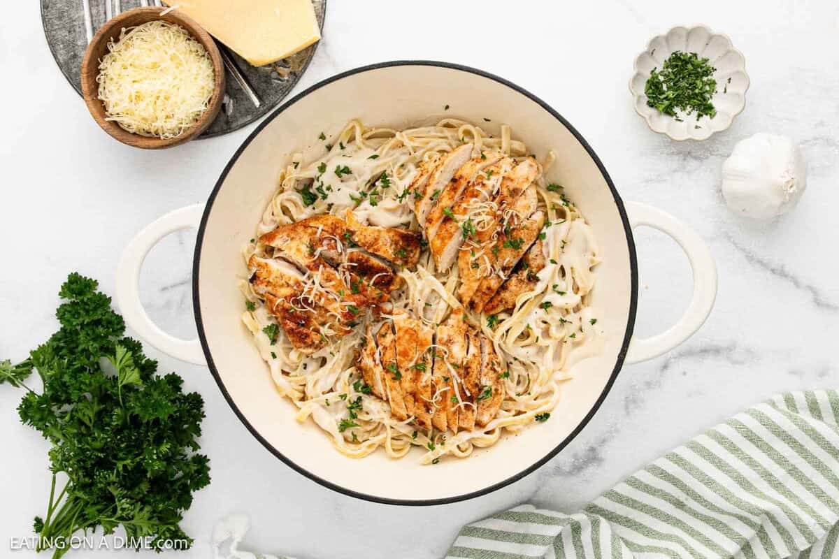 A white pot filled with creamy Chicken Fettuccine Alfredo, topped with sliced, seasoned chicken breast and sprinkled with chopped parsley and grated cheese. Fresh herbs, a garlic bulb, cheese, and a striped napkin sit nearby.