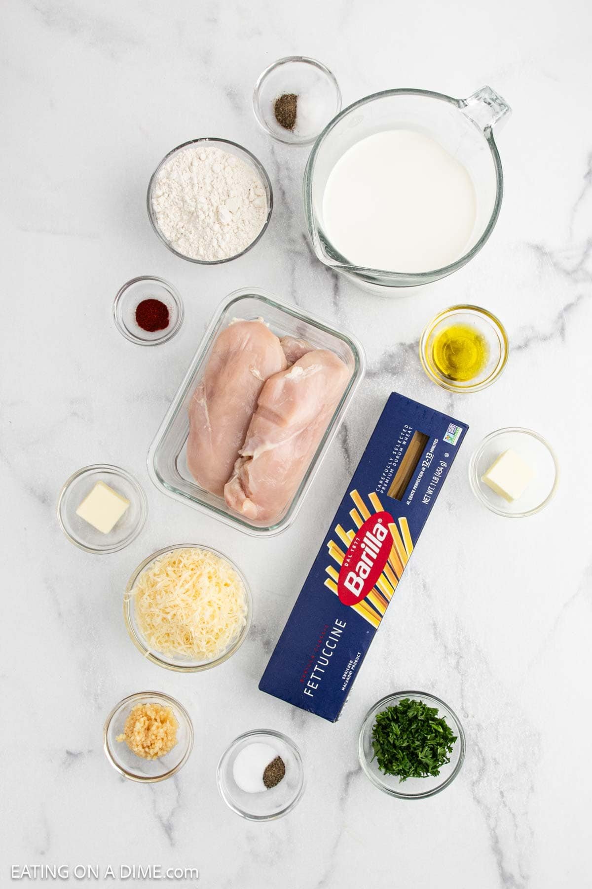 Overhead view of ingredients on a white surface for Chicken Fettuccine Alfredo, including raw chicken breasts, fettuccine pasta, shredded cheese, flour, milk, butter, oil, garlic, parsley, and small bowls of seasoning.